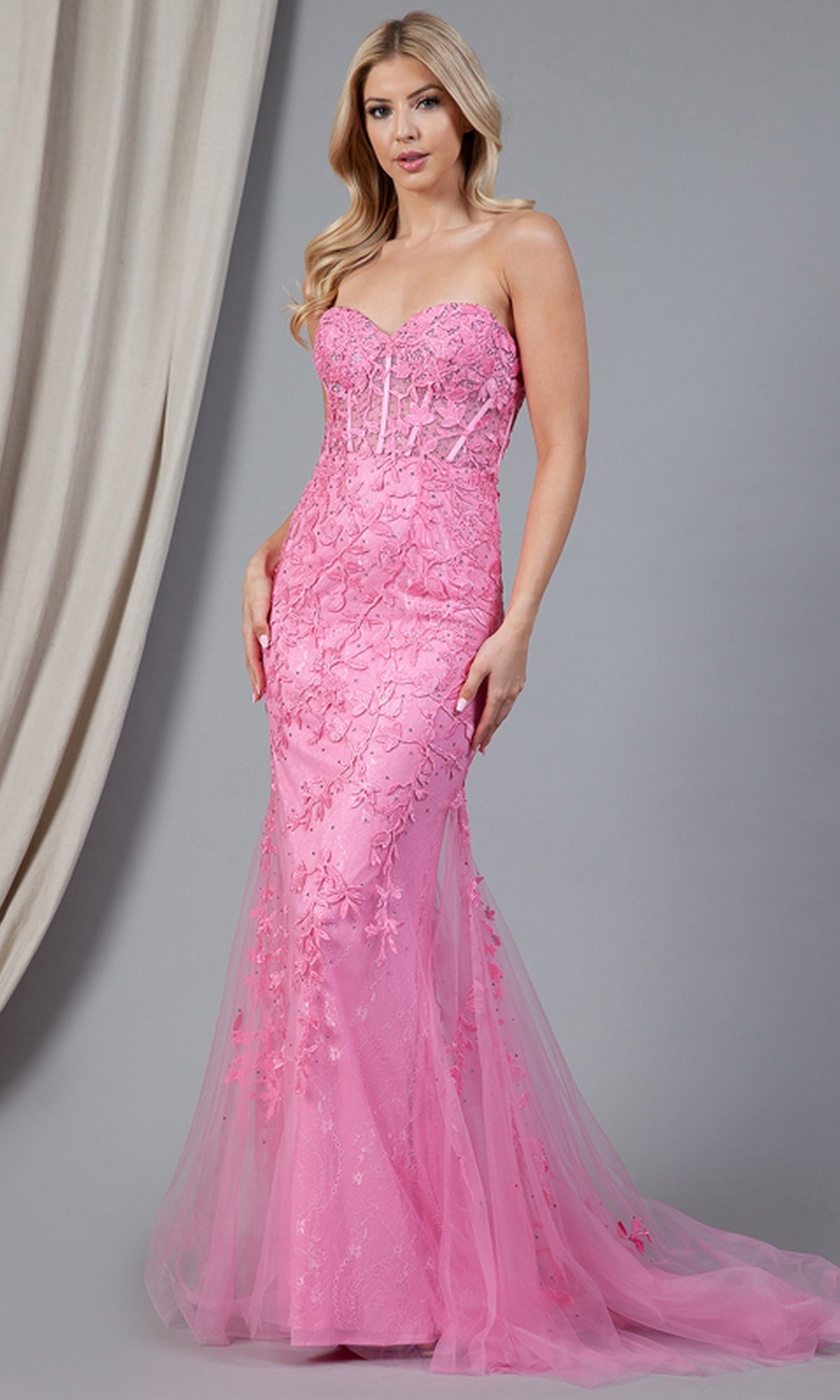 A woman wearing the Strapless Sweetheart Long Mermaid Prom Dress 7024, featuring a fitted pink mermaid silhouette with embroidered floral designs and sheer tulle overlay, poses against a gray background with draped curtains.